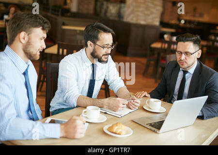Businessman in suit and glasses working in cafe showing ideas on laptop to two pleased colleagues in shirts and ties with tablet Stock Photo