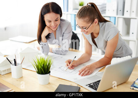Two young female office workers standing at desk bending over schemes of building, talking and smiling, business life Stock Photo
