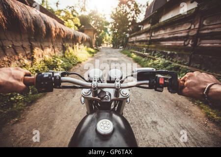 Biker driving his motorcycle on country road in a village. Point of view with focus is on the handlebar and man hands. Stock Photo