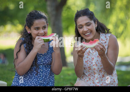Happy Hispanic mother with her teen daughter eating watermelon in park Stock Photo