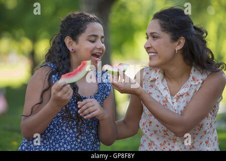 Happy Hispanic mother with her teen daughter eating watermelon in park Stock Photo