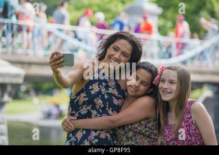 Happy Hispanic mother with two teen daughters taking selfie with mobile phone in park Stock Photo