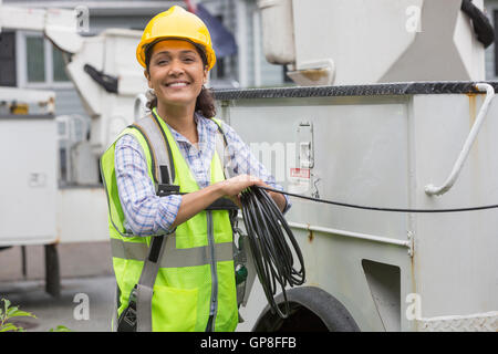 Portrait of happy Hispanic female utility worker holding cable by truck at site Stock Photo