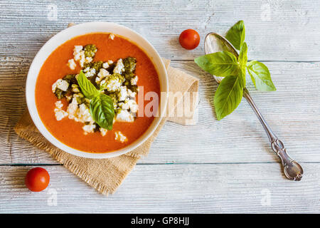 Homemade vegetarian tomato soup with feta cheese and pesto sauce in white bowl on wooden background. Served with fresh basil, to Stock Photo