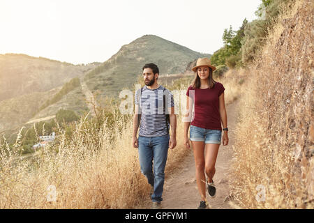 Nice young couple strolling down a mountain in very bright sunlight wearing t-shirts and jeans and a straw hat at sunset Stock Photo