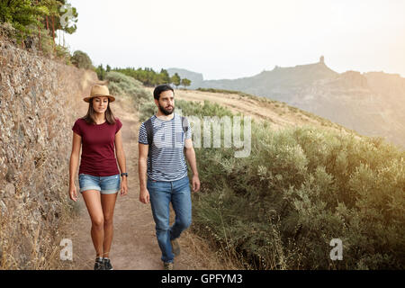 Cute young couple strolling down a mountain in bright sunshine wearing t-shirts and jeans and a straw hat at sunset Stock Photo