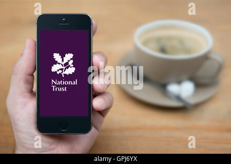 A man looks at his iPhone which displays the National Trust logo, while sat with a cup of coffee (Editorial use only). Stock Photo
