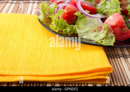 Detail of a transparent plate closeup with raw vegetable salad and a bright yellow napkin on a table covered with a bamboo mat Stock Photo