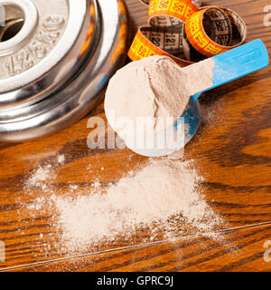 Whey protein powder in measuring scoop, meter tape and dumbbell on wooden background. Stock Photo