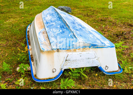 Upside down boat on land. The boat has blue keel and yellowing white sides. Dry leaves have started to accumulate around it. Stock Photo