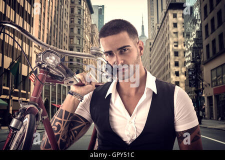 Portrait of young tattooed man in elegant clothes holding a bicycle in middle of a city street Stock Photo