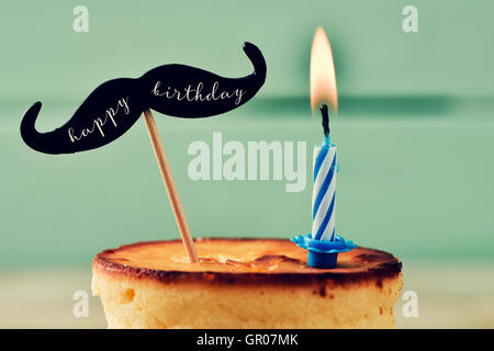 closeup of a cheesecake topped with a moustache with the text happy birthday written in it and attached to a stick, and a lit bi Stock Photo