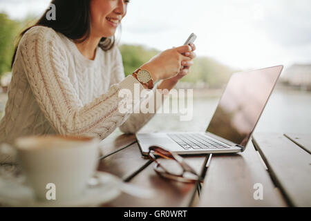 Cropped shot of young woman using mobile phone at outdoor cafe. Female sitting at table with laptop reading text message on her Stock Photo