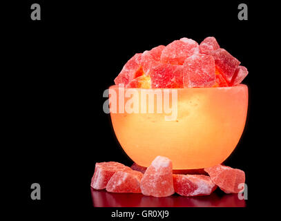 Turned on Himalayan pink salt lamp carved as a bowl on a black table with a black background. Stock Photo
