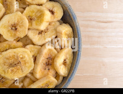 Top close view of dehydrated banana chips in an old stoneware bowl atop a wood table. Stock Photo