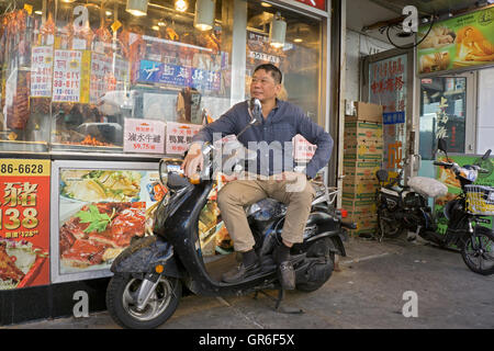 Portrait of an Asian American man sitting on a delivery person's scooter outside a restaurant in Downtown Flushing, Queens, NYC. Stock Photo