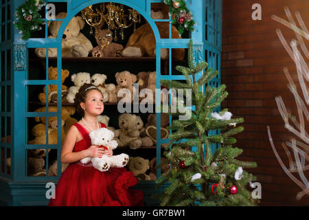 Adorable little girl, dressed in a lush red gown, hugging a plush bear next to the window with toys. Celebration Valentine's Day Stock Photo