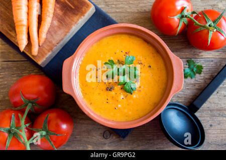 Vegetable tomato and carrot soup in ceramic pot and ingredients over rustic wooden background Stock Photo