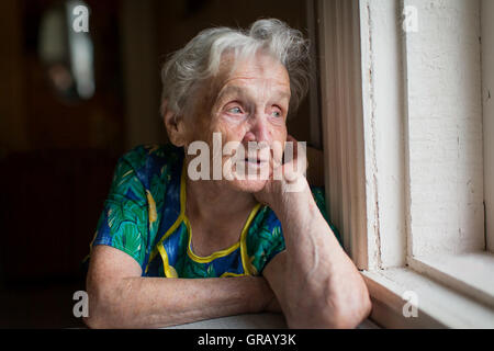 An elderly woman looks out the window sitting in the kitchen. Stock Photo