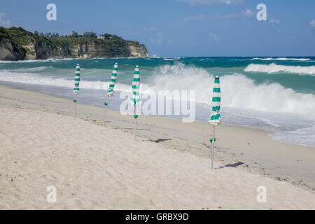 White Sandy Beach, Blue Sky, Waves, Closed Green And White Sun Shades, Cliffs With Some Trees In The Background. Tropea, Calabria, Italy Stock Photo