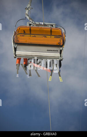 Single Chair From Chair Lift, Seen From Behind, With Orange Weather Protectors, Legs With Skis Against The Blue Sky And White Clouds Stock Photo