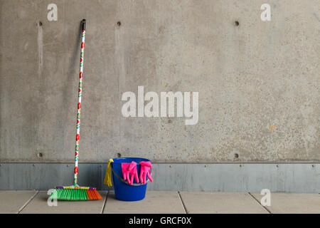 Colorful cleaning equipment (broom, bucket, rag and gloves) in front of a gray, monotonous concrete wall and stained floor. Stock Photo