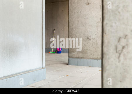 Colorful cleaning equipment (broom, bucket, rag and gloves) in front of a gray, monotonous concrete wall and stained floor. Stock Photo