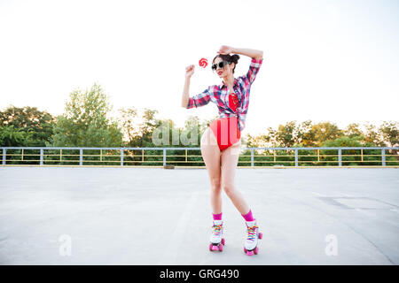 Beautiful young woman in red swimsuit and plaid shirt holding lollipop and standing on roller skates Stock Photo
