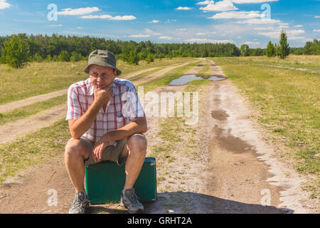 Mature man with old suitcase sitting on an country road and thinking Stock Photo