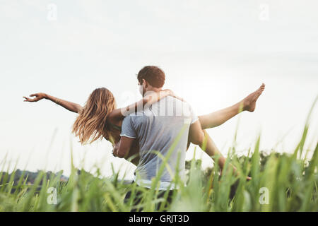 Rear view shot of man carrying woman in rural field. Couple enjoying outdoors on grass field. Stock Photo