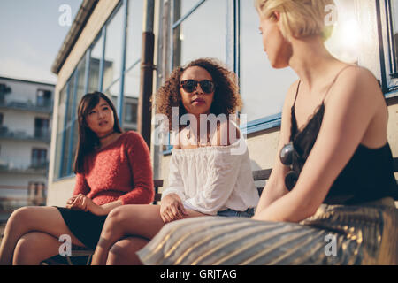 Three young female friends meeting outdoors. Multiracial group of young women sitting outdoors and chatting. Stock Photo