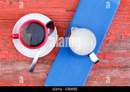 Freshly brewed cup of black coffee in a red mug on a white saucer served with a jug of milk and blue napkin on a grunge wooden r Stock Photo