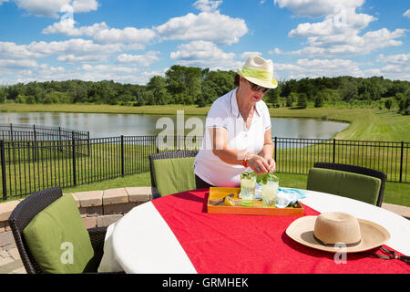 Senior lady preparing iced drinks outdoors adding fresh mint and lemon to glasses on a tray on a garden table with red cloth ove Stock Photo