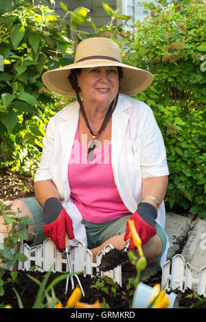 Smiling active elderly lady tending flowerbeds around the house sitting in her wide brimmed sunhat on the pathway paving weeding Stock Photo