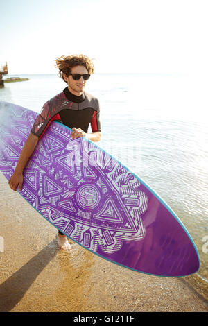 Happy attractive young man surfer in sunglasses with surfing board running on the beach Stock Photo