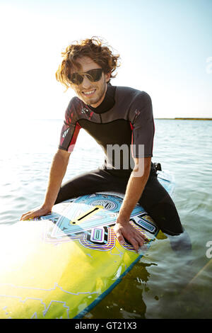 Smiling young male surfer sitting on his surf board in water wearing swimsuit and looking at camera Stock Photo