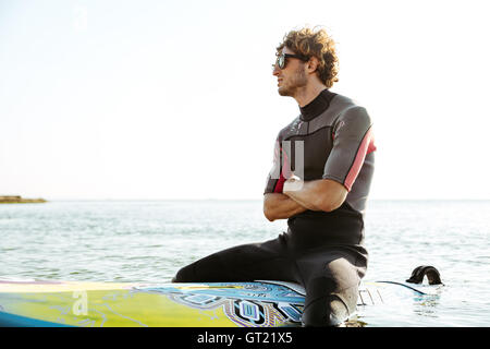 Smiling young male surfer sitting on his surf board with hands crossed in ocean wearing swimsuit Stock Photo