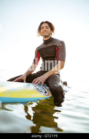 Smiling young male surfer sitting on his surf board in water wearing swimsuit and looking at camera Stock Photo