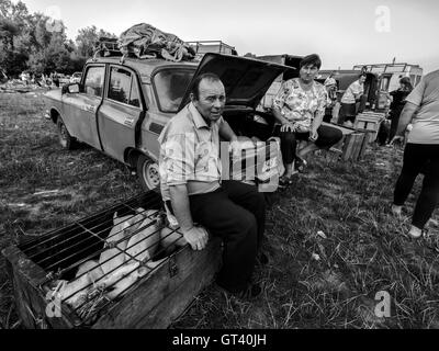 Middle-aged man sitting in a cage with piglets, his wife standing beside the car, in the trunk where they brought pets for sale  Stock Photo