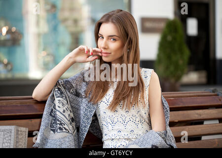 Cute young brunette woman sitting on the bench in summer street Stock Photo