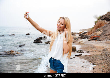 Young smiling beautiful woman listening music and making selfie while standing on the rocky beach Stock Photo