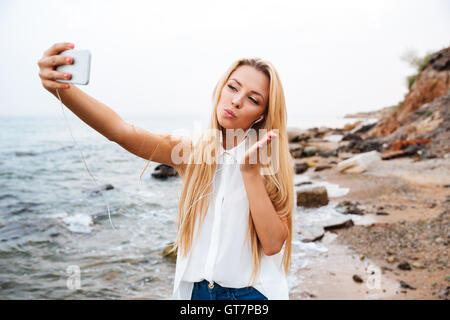 Young smiling beautiful woman sending air kiss and making selfie while standing on the rocky beach Stock Photo