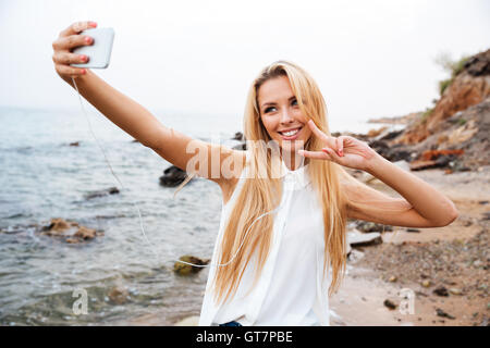 Young smiling beautiful woman showing v gesture and making selfie while standing on the rocky beach Stock Photo