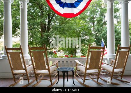 Four vacant wooden rocking chairs lined up on a patio overlooking a lush garden below a draped American flag symbolising 4th Jul Stock Photo