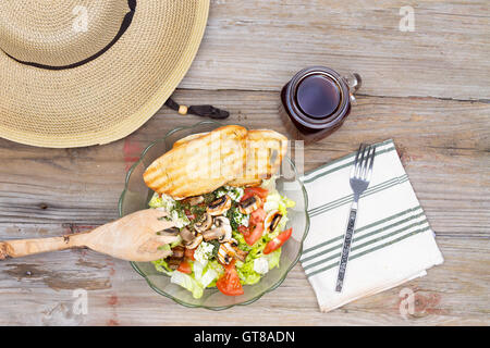 Wide-Brimmed Straw Hat and Healthy Lunch of Salad and Bread with Black Tea on Wooden Background as seen from Above Stock Photo
