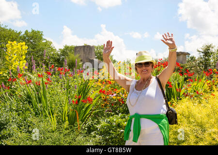 Trendy joyful Grandma outdoors in her garden laughing and waving her hands in the air in front of a bed of colorful summer flowe Stock Photo