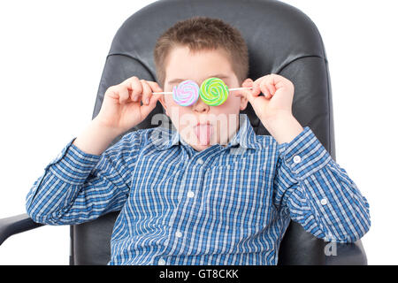 Close up Funny White Boy in Checkered Shirt, Sitting on an Office Chair and Holding Lollipops Over his Two Eyes with Tongue Out. Stock Photo