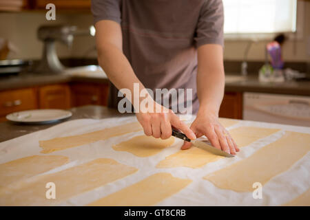 Hands of a man preparing fresh homemade fettuccine pasta trimming the sheets of rolled dough into rectangles to feed through the Stock Photo