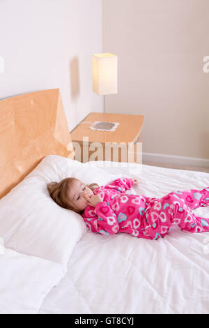 Adorable small girl in pink pajamas lying resting on a big king size bed picking her nose as she awakens from her daily nap Stock Photo