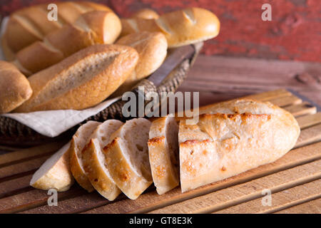 Sliced fresh baguette on a cutting board with a basket of crusty golden rolls and buns on display at a buffet or bakery, close u Stock Photo